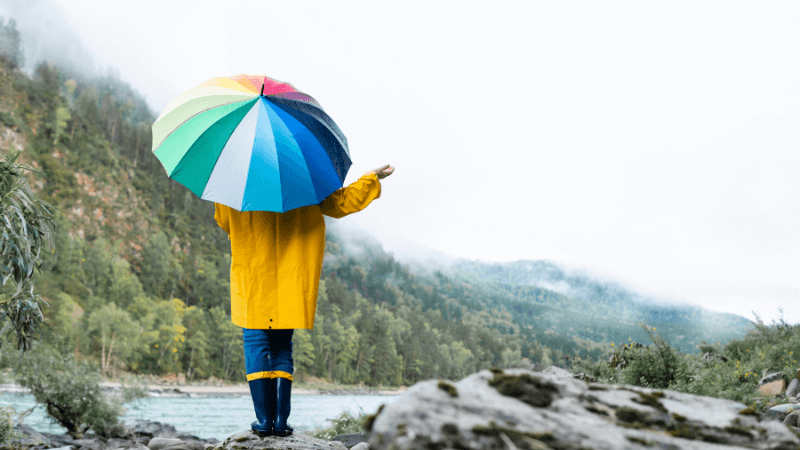 A person in a yellow rain coat with a rainbow umbrella feels for rain while standing on rocks near water