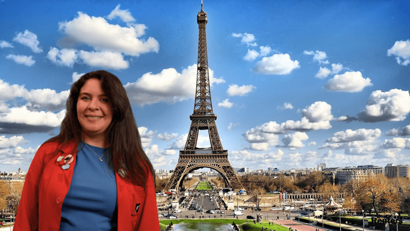 Museum Educator Ana in a red lab coat smiling in front of the Eiffel Tower. 