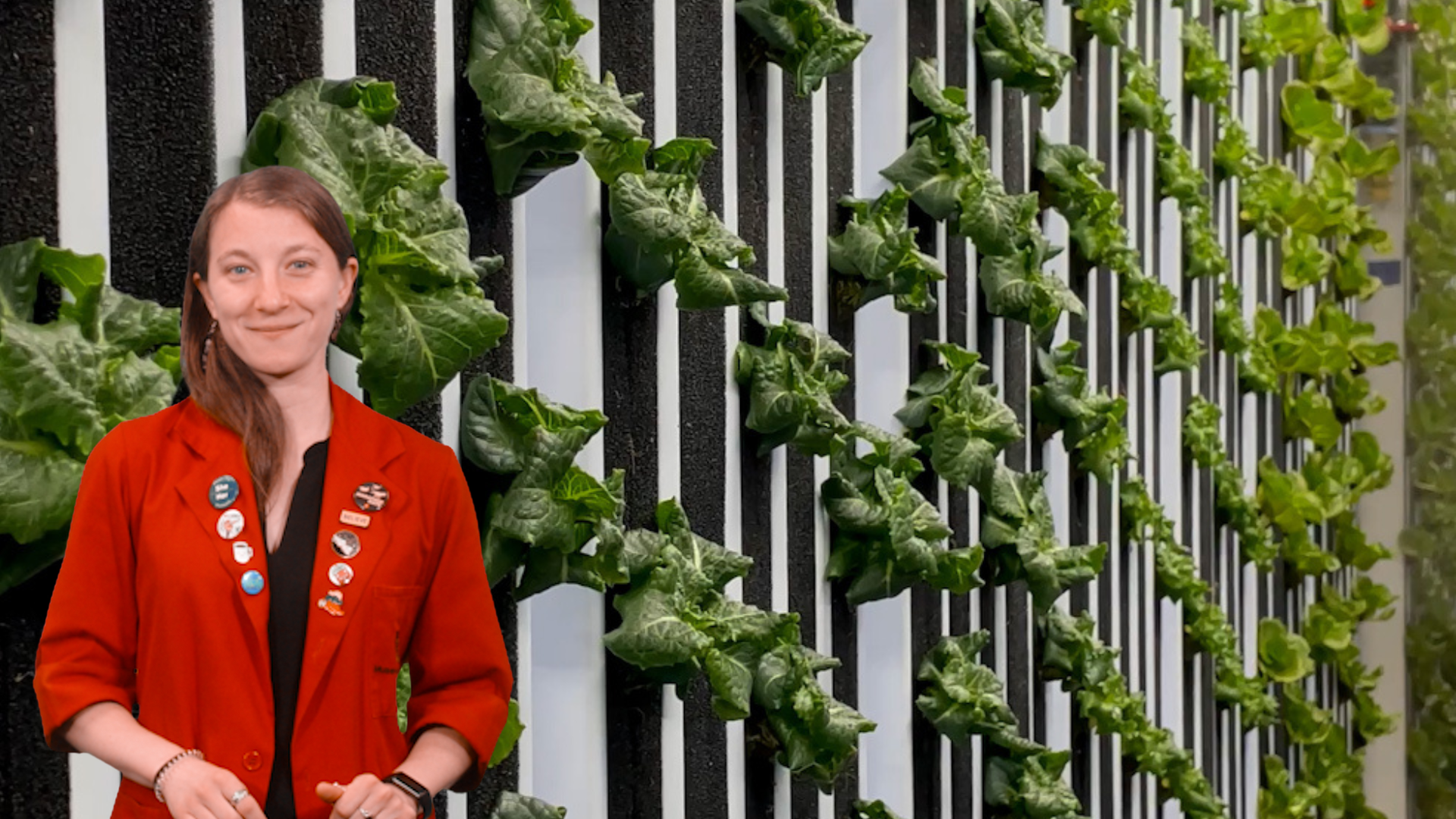 Museum Educator Sloane in a red lab coat in front of a hydroponic farming system. 