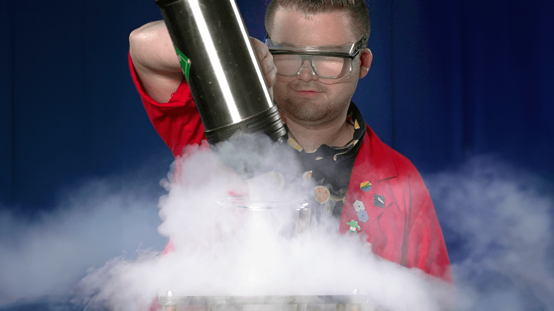 Museum Educator Locke wearing a red lab coat and safety glasses pouring liquid nitrogen into a bucket.