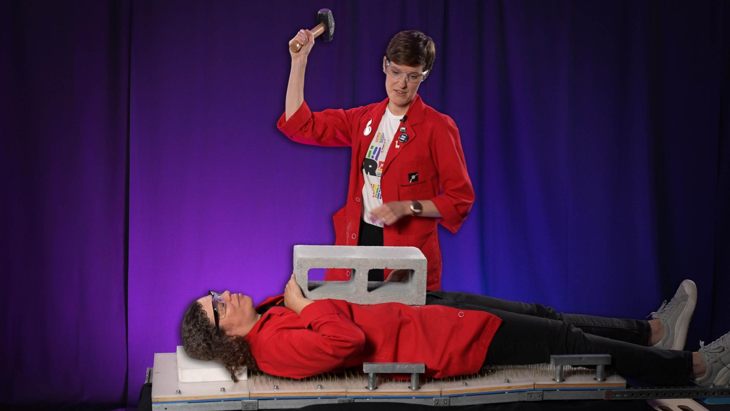 Emily holding a sledgehammer over Jeannine on a bed of nails with a cinder block on her chest