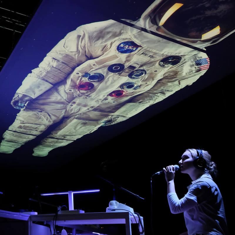 A performer hold a microphone with a space suite projected on the ceiling above them.