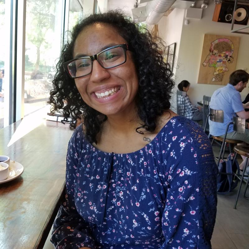 Valerie Novack: Birracial woman with curly, shoulder-length black hair and glasses is smiling while seated in a shop. She is wearing a blue flowered top.