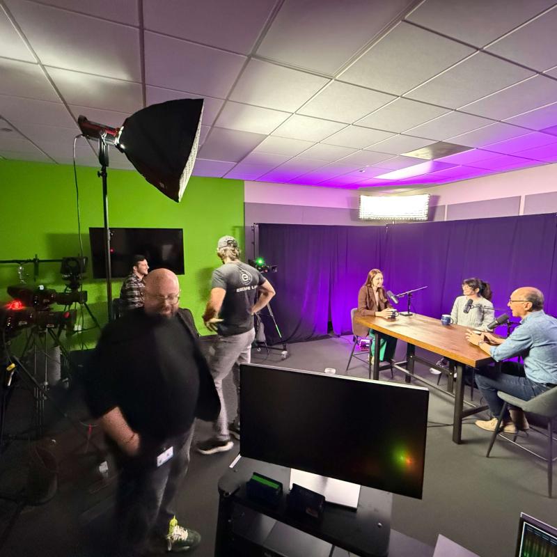 A view of a video studio with three people sitting at a table in front of a curtain and a crew working with cameras